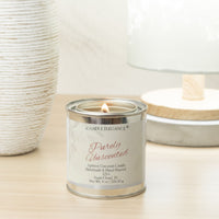 A lit candle in a silver metal tin with a white label reading 'Candle Elegance Purely Unscented,' placed on a light wood surface with decorative home decor in the background.