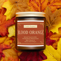 A jar candle labeled 'Blood Orange' by Candle Elegance, placed on a bed of colorful autumn leaves. The candle has a brown label with gold text and a gold metal lid.