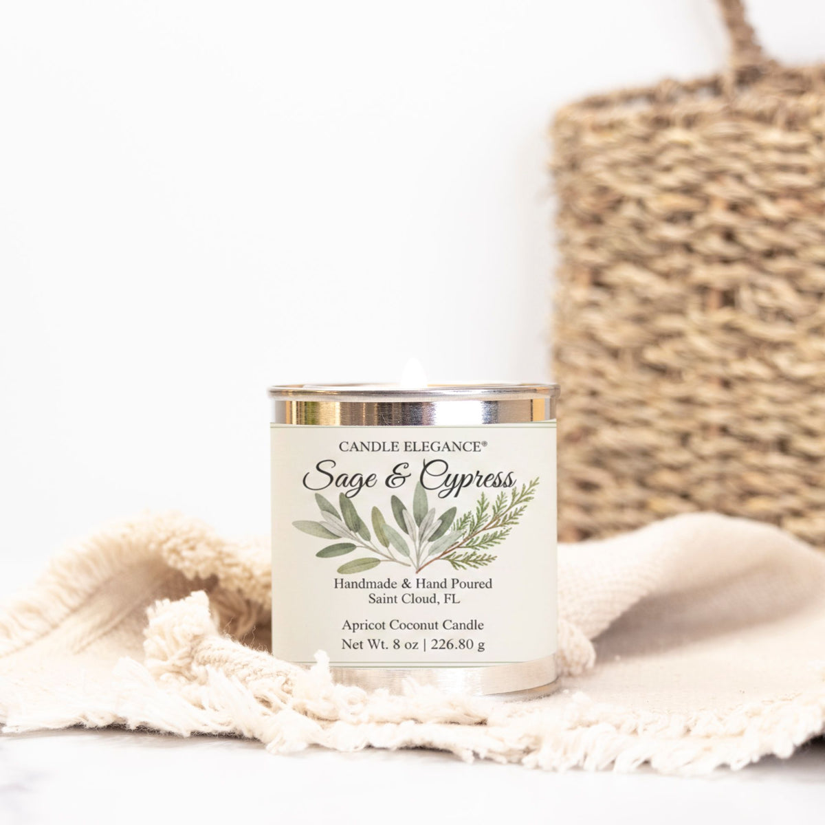 A jar candle labeled 'Candle Elegance Sage & Cypress' sits on a cream fringed cloth with a woven basket blurred in the background.