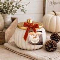 A white ribbed pumpkin-shaped candle jar with a gold lid and a rust velvet bow, decorated with a round product label. The candle is styled on a beige cloth with pinecones, a wooden tray, and another white pumpkin in the background.