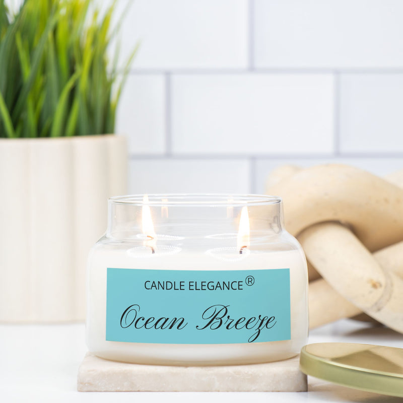 A jar candle with white wax, a gold metal lid, and a light blue label reading 'CANDLE ELEGANCE' and 'Ocean Breeze' on a light gray background.