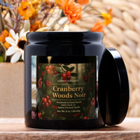 A black glass jar candle with its lid leaning against it on a wooden surface. The front label reads 'Candle Elegance Cranberry Woods Noir' with an illustration of red cranberries on a dark background.