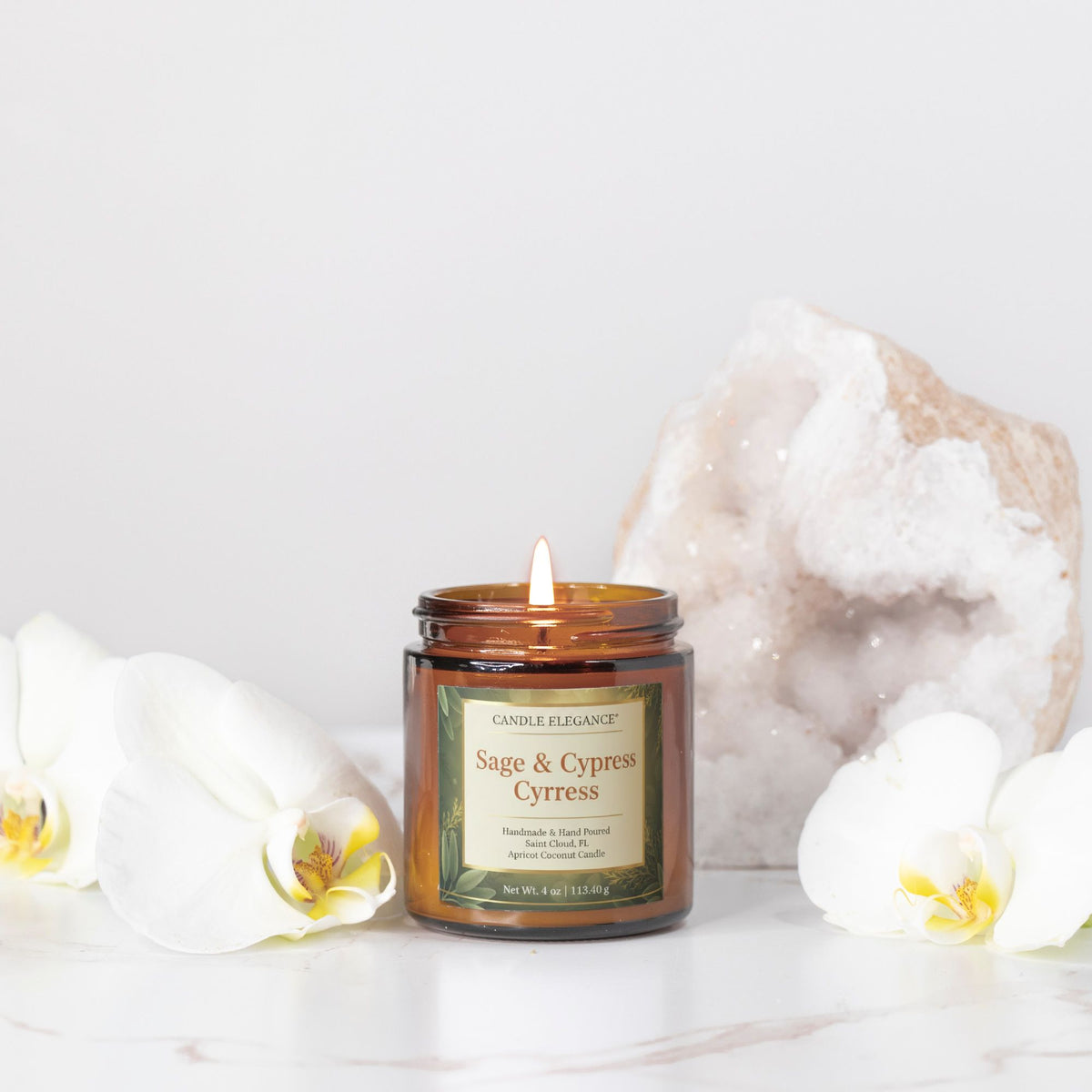 An amber glass jar candle labeled 'Candle Elegance Sage & Cypress' with a lit wick, styled on a white marble surface with white orchids and a large geode crystal in the background.