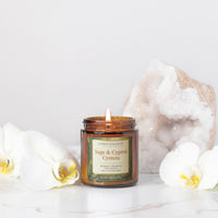 An amber glass jar candle labeled 'Candle Elegance Sage & Cypress' with a lit wick, styled on a white marble surface with white orchids and a large geode crystal in the background.