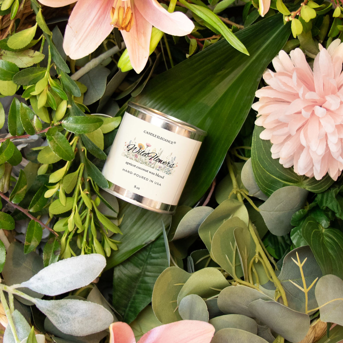 A Wildflowers candle tin with a white label reading 'Wildflower' and 'Hand-poured in San Diego' is nestled among green leaves, pink flowers, and eucalyptus in a styled flat-lay arrangement.