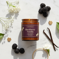 An amber glass jar candle labeled Candle Elegance Blackberry Vanilla, styled on a white marble surface with blackberries, vanilla beans, white flowers, and small gold hexagonal accents.