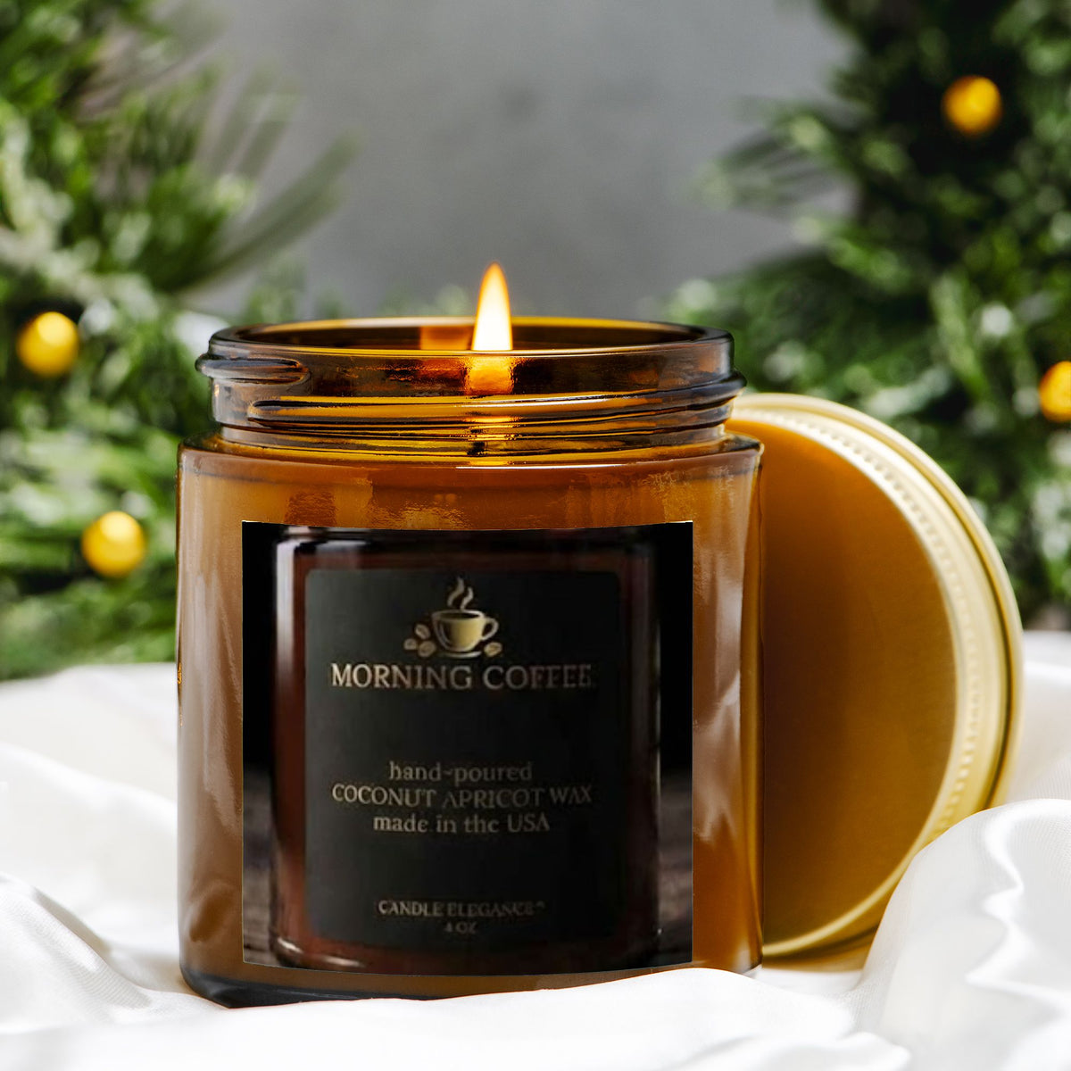 A lit amber glass candle jar labeled 'MORNING COFFEE' with a gold lid resting beside it, set on white fabric with blurred greenery and gold ornaments in the background.