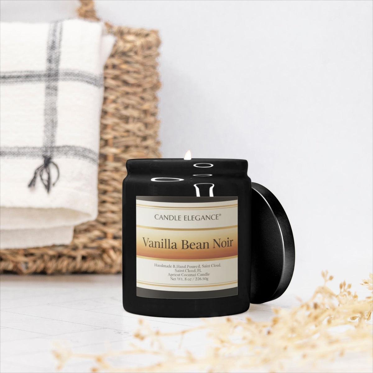 A black glass jar candle with a white label reading 'Pink Watermelon Lemonade Noir' by Candle Elegance, shown with its lid leaning beside it on a light surface. A woven basket and folded white towel are blurred in the background.