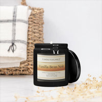 A black glass jar candle with a white label reading 'Pink Watermelon Lemonade Noir' by Candle Elegance, shown with its lid leaning beside it on a light surface. A woven basket and folded white towel are blurred in the background.