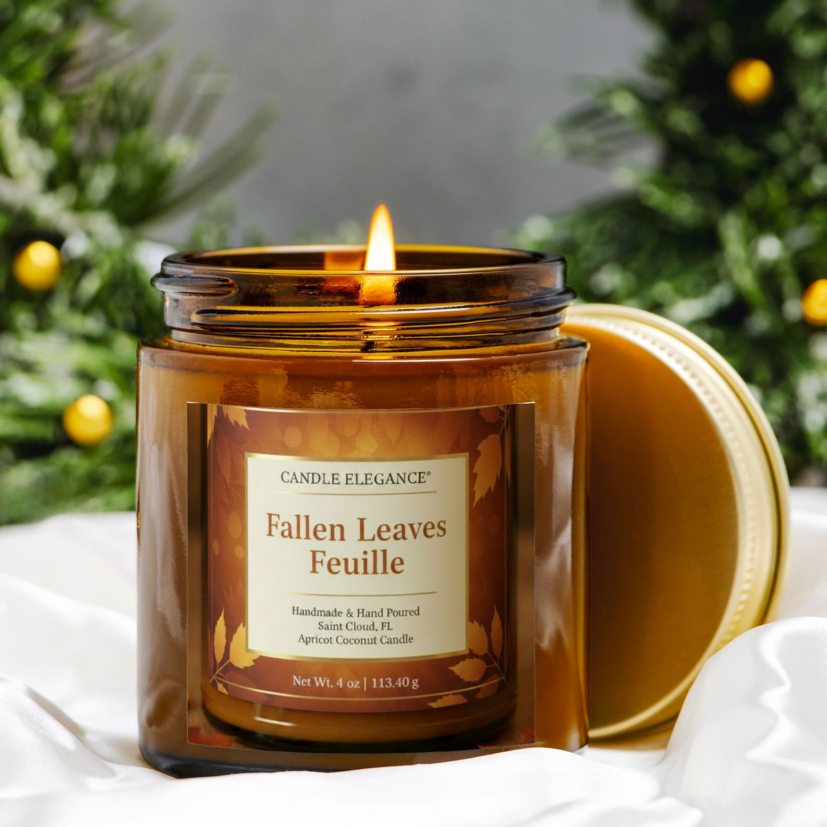 A lit amber glass jar candle with a gold lid resting beside it on white fabric, set against a blurred holiday background with greenery and gold ornaments. The front label reads Candle Elegance and Fallen Leaves Feuille.