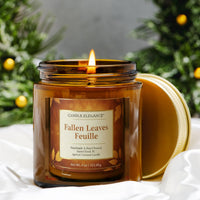 A lit amber glass jar candle with a gold lid resting beside it on white fabric, set against a blurred holiday background with greenery and gold ornaments. The front label reads Candle Elegance and Fallen Leaves Feuille.