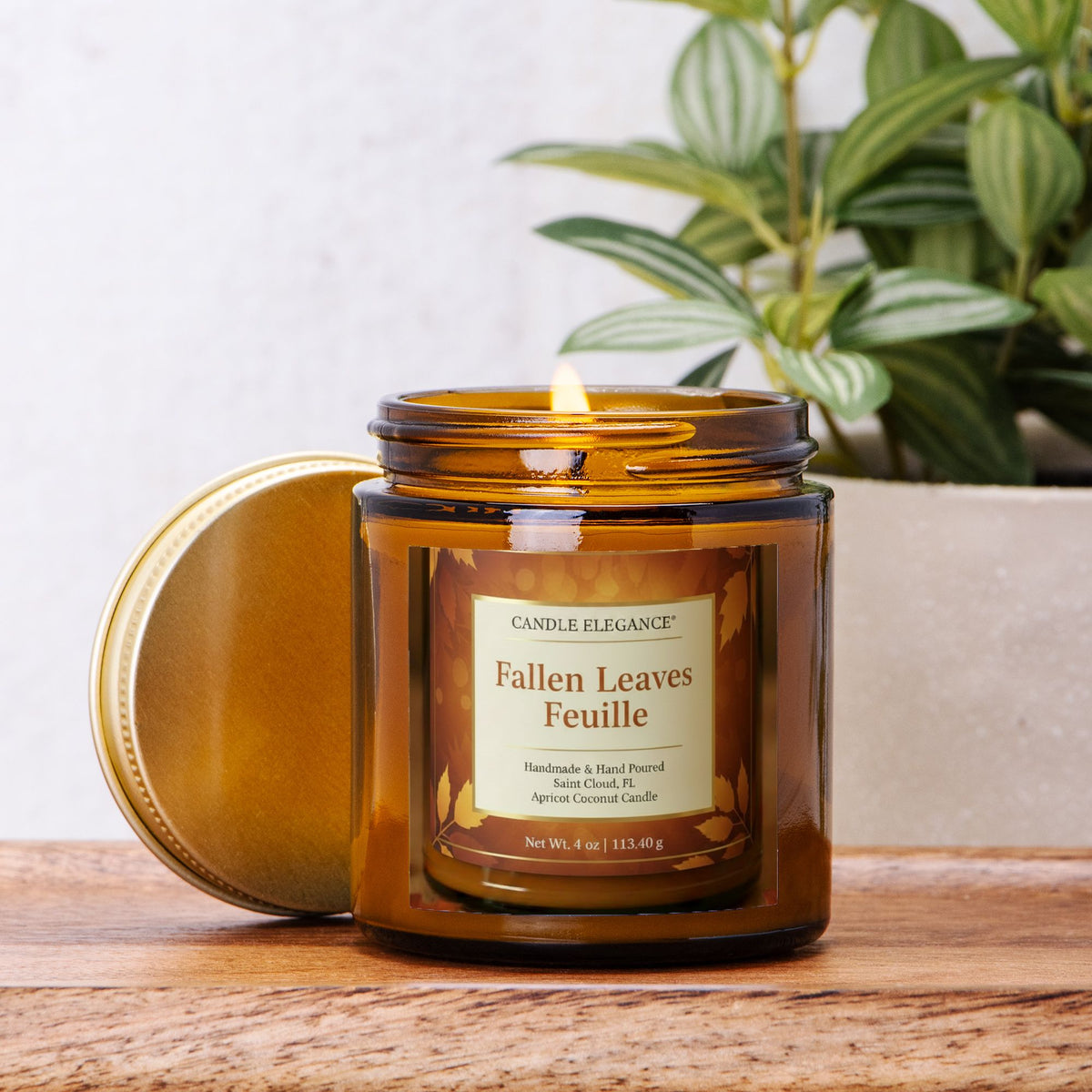 An amber glass jar candle with its lid leaning beside it on a wooden surface. The front label reads 'Candle Elegance' and 'Fallen Leaves Feuille.