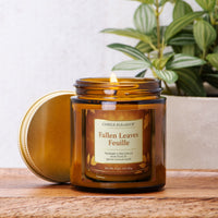 An amber glass jar candle with its lid leaning beside it on a wooden surface. The front label reads 'Candle Elegance' and 'Fallen Leaves Feuille.