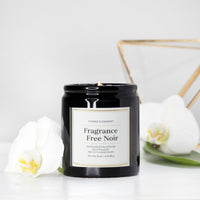 A black glass jar candle labeled 'Fragrance Free Noir' by Candle Elegance, styled with white orchids and a gold geometric frame on a white surface against a white background.