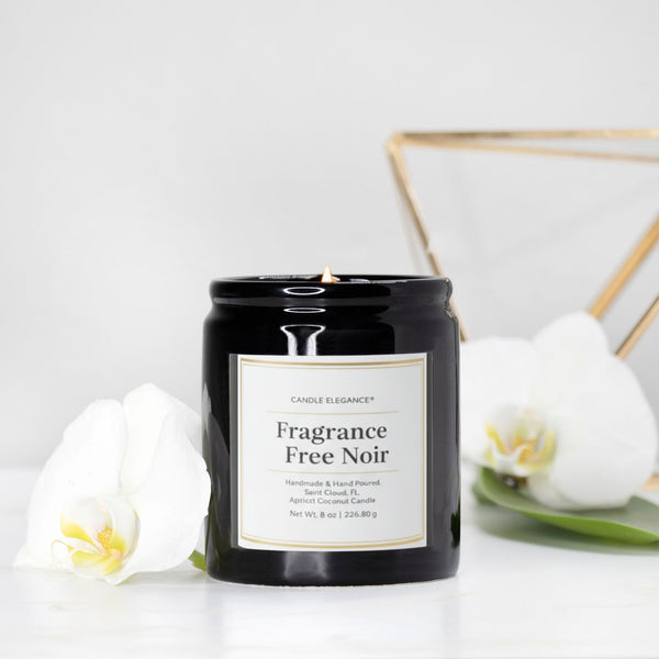 A black glass jar candle labeled 'Fragrance Free Noir' by Candle Elegance, styled with white orchids and a gold geometric frame on a white surface against a white background.