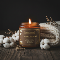 A lit amber glass jar candle labeled 'Candle Elegance Cashmere Musk' sits on a rustic wooden surface, surrounded by cotton bolls and a folded cream knit blanket against a dark background.
