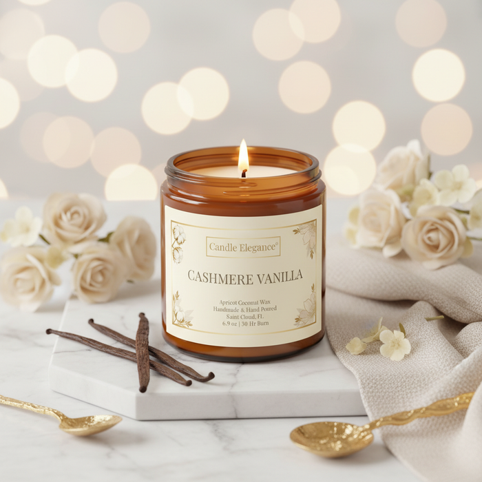 A lit amber glass jar candle labeled 'Candle Elegance Cashmere Vanilla' sits on a white marble board with vanilla beans, cream flowers, a beige cloth, and gold spoons against a soft bokeh background.