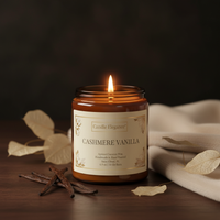 A lit amber glass jar candle labeled 'Candle Elegancee' in 'Cashmere Vanilla' scent, styled on a dark wooden surface with vanilla beans, dried leaves, and a cream fabric runner.
