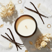 A single-wick candle in a dark amber glass jar sits on a white marble surface, surrounded by vanilla beans, cream flowers, feathers, and small gold leaves. The candle wax is a warm off-white, and the jar has a dark brown rim.