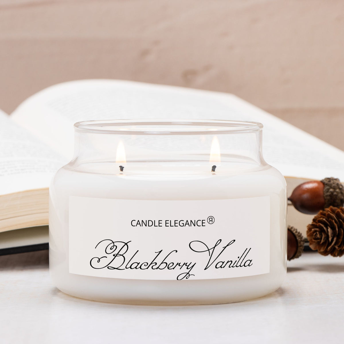 A double-wick candle in a clear glass jar with white wax, labeled 'Candle Elegance Blackberry Vanilla,' styled on a light surface with an open book and acorns in the background.