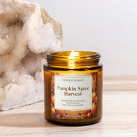 A lit amber glass jar candle labeled 'Pumpkin Spice Harvest' by Candle Elegance, placed on a light wood surface with a large white crystal cluster in the background.