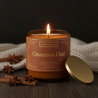 A lit Candle Elegance Cinnamon Chai scented candle in a clear glass jar with a warm brown label, shown on a dark wooden surface with a gold lid leaning beside it and cinnamon sticks and star anise nearby.