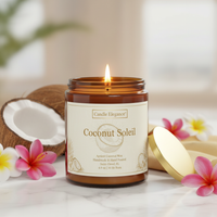 A lit amber glass jar candle labeled 'Coconut Soleil' by Candle Elegance, placed on a marble surface with a coconut half, pink flowers, and a gold lid nearby.