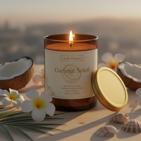 A lit amber glass candle with a cream label reading 'Coconut Soleil' by Candle Elegance, shown on a wooden surface with a gold lid beside it, surrounded by white plumeria flowers, coconut halves, and seashells against a softly blurred beach background.