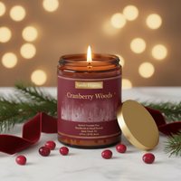 A lit amber glass jar candle labeled 'Cranberry Woods' by Candle Elegance, with its gold lid resting beside it on a marble surface. Pine branches, red ribbon, and scattered cranberries decorate the scene against a warm blurred bokeh background.