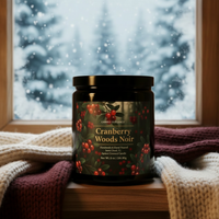 A black glass jar candle labeled 'Candle Elegance Cranberry Woods Noir' sits on a wooden surface with scarfs around it against a snowy winter scene window backdrop.