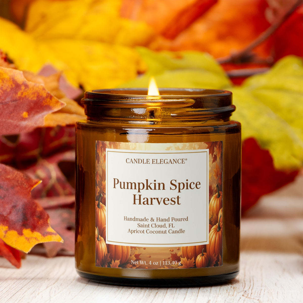 A lit amber glass jar candle labeled 'Pumpkin Spice Harvest' by Candle Elegance, set on a light wood surface with autumn leaves in the background.