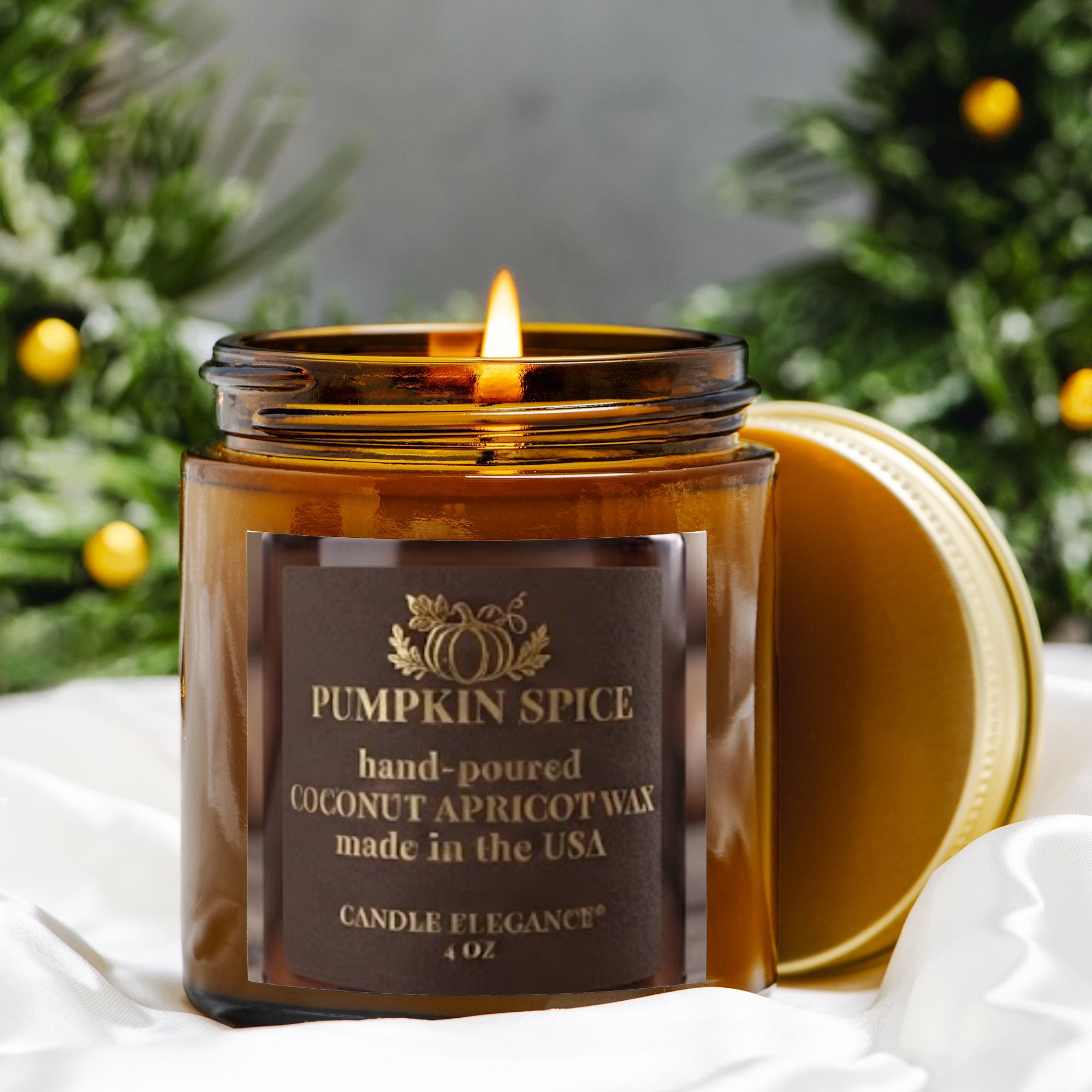 A lit amber glass jar candle labeled 'Pumpkin Spice' by Candle Elegance, with its gold lid resting beside it on white fabric. The background features blurred green pine branches with small yellow ornaments.
