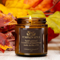 A lit amber glass jar candle labeled 'Pumpkin Spice' by Candle by Elegance, set against a background of colorful autumn leaves on a light wood surface.