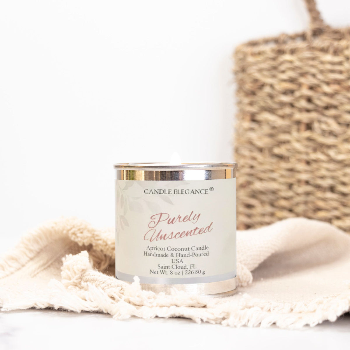 A Candle Elegance 'Purely Unscented' apricot coconut candle in a clear glass jar with a white label and gold-toned lid, styled on a cream fringed cloth with a woven basket in the background.