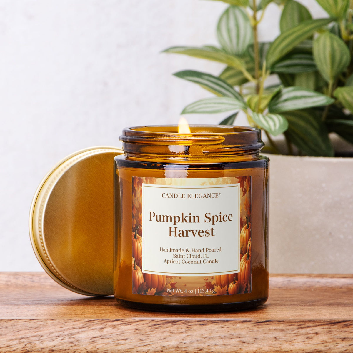 An amber glass jar candle labeled 'Pumpkin Spice Harvest' by Candle Elegance, with its gold lid resting beside it on a wooden surface. A green potted plant is visible in the softly blurred background.