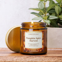 An amber glass jar candle labeled 'Pumpkin Spice Harvest' by Candle Elegance, with its gold lid resting beside it on a wooden surface. A green potted plant is visible in the softly blurred background.