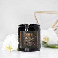 A black glass jar candle labeled 'Blackberry Vanilla Noir' sits on a white surface with white orchid flowers and a gold geometric frame in the background.