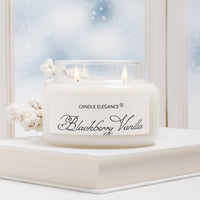 A lit double-wick candle in a clear glass jar with cream-colored wax, labeled 'Candle Elegance Blackberry Vanilla,' styled on top of a white book with soft white decorative berries against a snowy window background.