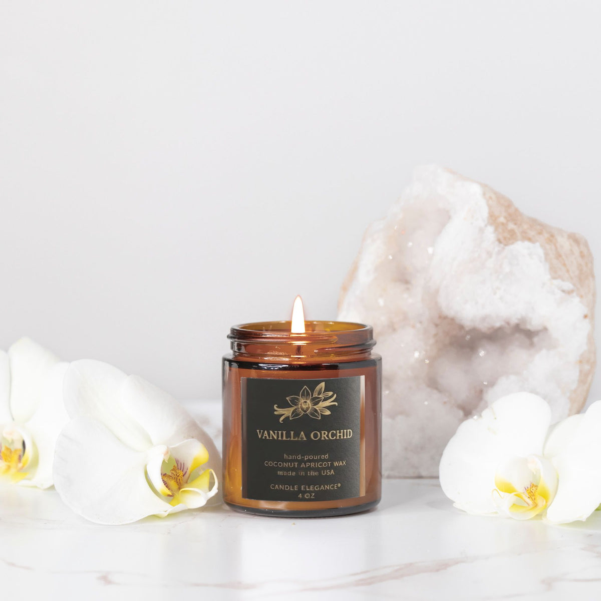 An amber glass jar candle labeled 'Vanilla Orchid' with a lit wick, styled on a white marble surface with white orchid flowers and a large white geode crystal in the background.