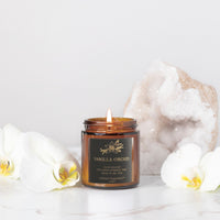 An amber glass jar candle labeled 'Vanilla Orchid' with a lit wick, styled on a white marble surface with white orchid flowers and a large white geode crystal in the background.