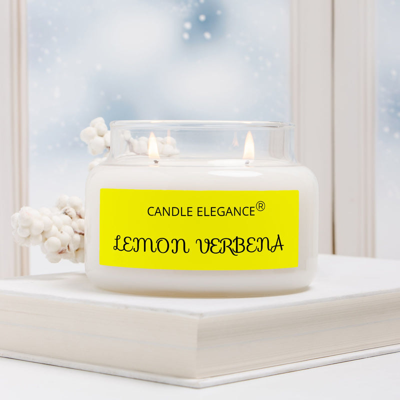 A double-wick candle in a clear glass jar with white wax sits on a white book. The jar has a bright yellow label reading 'CANDLE ELEGANCE Lemon Verbena.