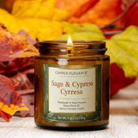A lit amber glass jar candle labeled 'Candle Elegance Sage & Cypress' sits on a light wood surface with autumn leaves in the background.