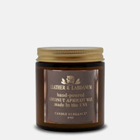 A 4 oz amber glass jar candle with a gold metal lid and a brown label reading 'Leather & Labdanum' and 'Coconut Apricot Wax'.