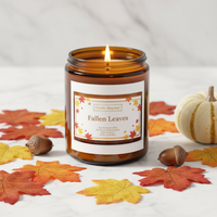 An amber glass jar candle labeled 'Fallen Leaves' by Candle Beguine, lit and styled on a white marble surface with autumn leaves, acorns, and a small white pumpkin.