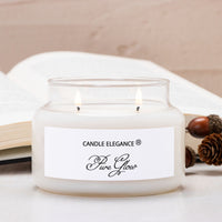A lit double-wick candle in a frosted glass jar with a white label reading 'CANDLE ELEGANCE' and 'Fire Glow', styled on a tabletop with an open book and acorns in the background.