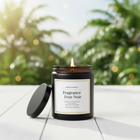 A lit candle in a dark amber glass jar sits on a white wooden surface with its black lid leaning beside it. The background is a softly blurred outdoor scene with green foliage and warm bokeh lights.