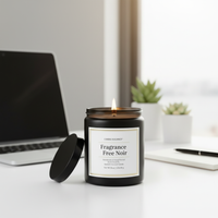 A lit black candle jar with its lid off, placed on a white desk next to a laptop, with small potted plants and a notebook in the background. The label reads 'Candle Island' and 'Fragrance Free Noir'.