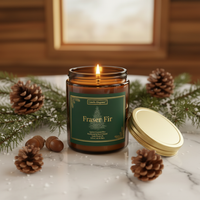 A lit amber glass jar candle labeled 'Fraser Fir' by Candle Emperor, styled on a marble surface with pine branches, pinecones, and acorns in front of a warm wood-paneled background.