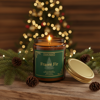 A lit amber glass jar candle labeled 'Fraser Fir' by Candle Dispo, set on a wooden surface with pine branches, pinecones, and a blurred Christmas tree in the background.