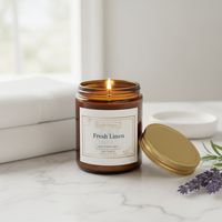 An amber glass jar candle labeled 'Fresh Linen' by Candle Equator, shown lit on a marble surface with its gold lid beside it, white towels, and lavender sprigs in a softly lit room.